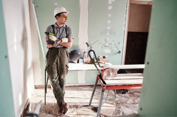 Construction worker in a helmet standing in a partially renovated room with tools, exposed walls, and construction equipment.