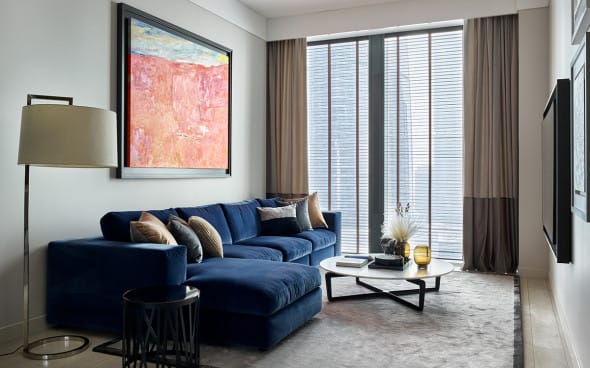 Modern living room with a deep blue velvet sectional sofa, abstract artwork, floor lamp, and tall windows with neutral curtains.